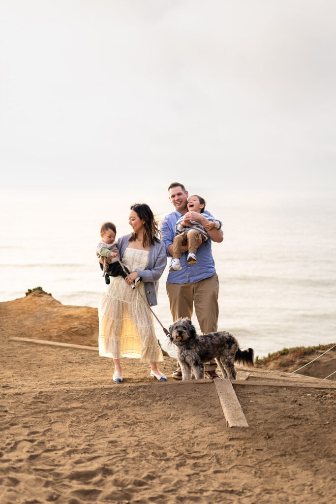 Parents walk along a sandy coastal path holding their children as their dog walks ahead, guided gently during a well led photo experience.
