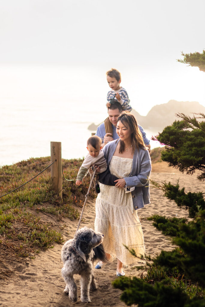 A father carries his toddler on his shoulders while the mother walks beside him holding the baby as they move along a sandy trail.