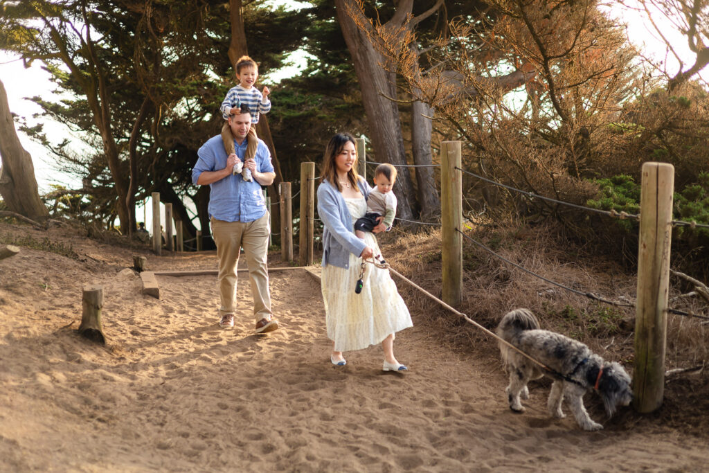 A mother leads the family dog on a leash while holding her baby as the father follows behind carrying their toddler on his shoulders.