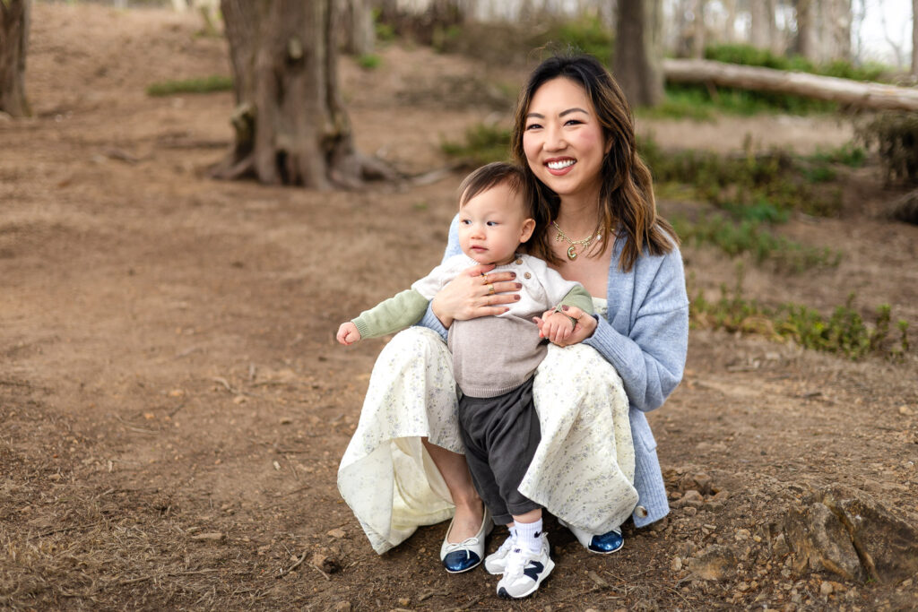 A mother crouches beside her baby, smiling toward the camera while holding her child comfortably in a relaxed outdoor environment.