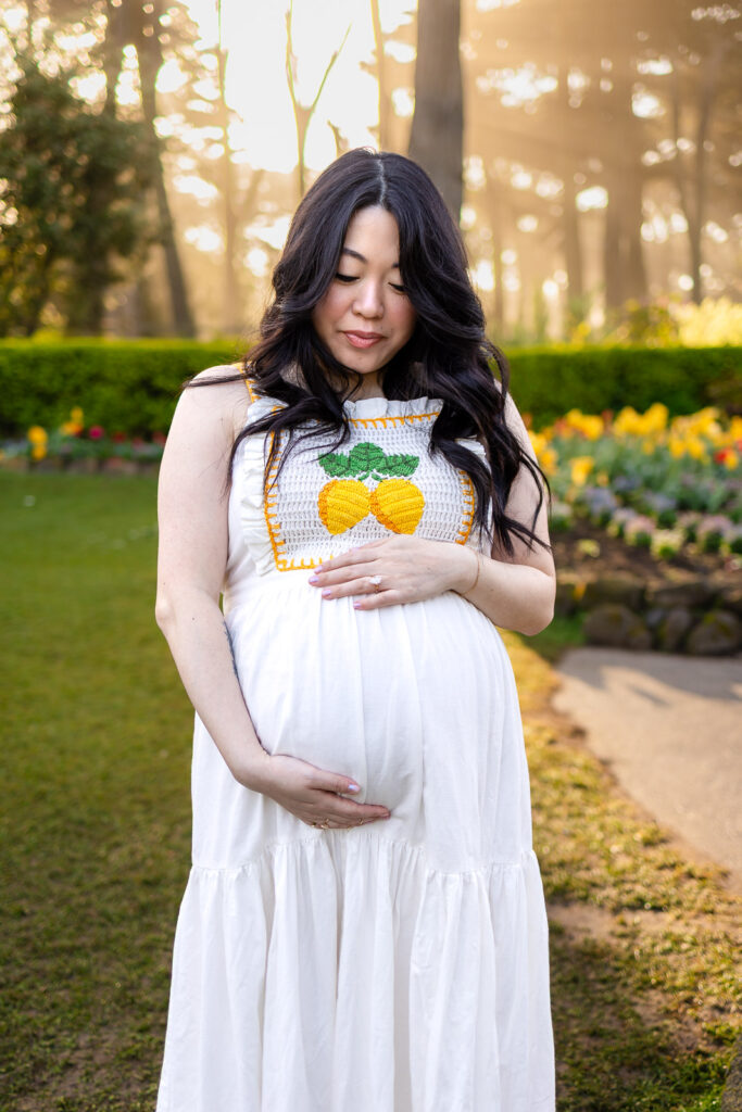 A mother in a long white dress holds her baby bump, illuminated by the soft light of golden hour during a spring photography session in San Francisco.