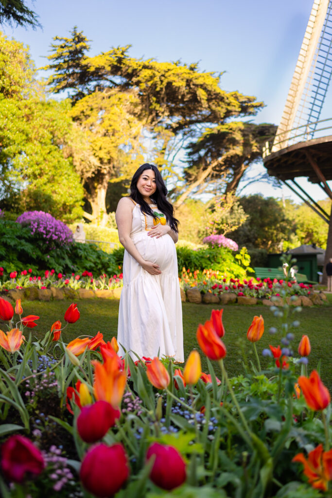 A serene maternity portrait of an expectant mother in a white dress, framed by colorful blooming tulips with the Golden Gate Park windmill in the background.