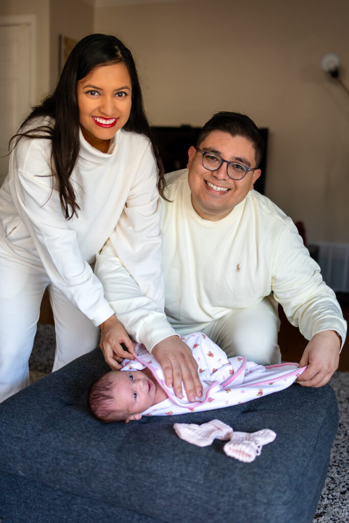 Parents dressed in soft neutral clothing lean over their swaddled newborn resting on a gray ottoman, smiling warmly toward the camera in their living room.