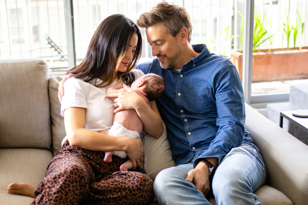 Parents sit close together on a couch, gazing at their newborn resting against the mother’s chest in a bright, modern living space.