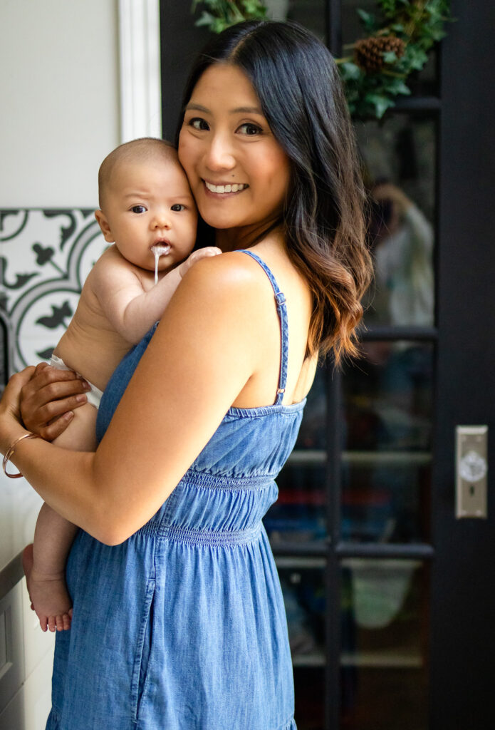 Mother holds her baby over her shoulder near a glass door, both looking toward the camera with natural window light illuminating their faces.