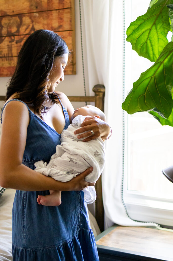 Mother cradles her swaddled newborn near a window with soft curtains and indoor plants, using the Best Light for Newborn Photos in San Mateo County Homes to create gentle shadows and clean skin tones.