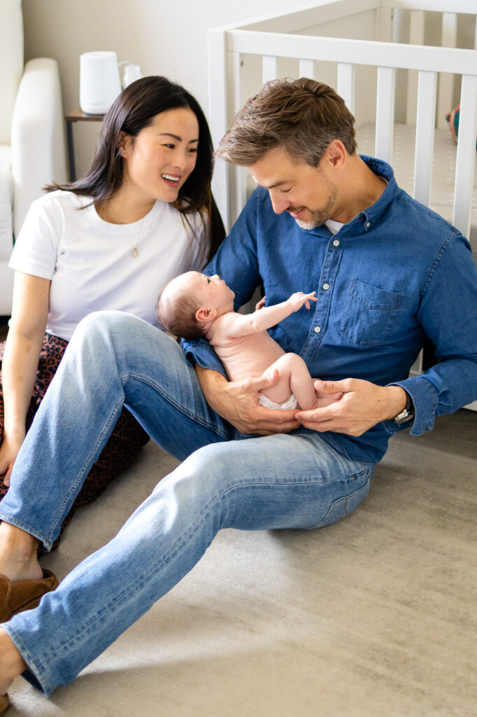 Parents sit on the floor beside a crib, smiling down at their diapered newborn resting comfortably in the father’s arms.