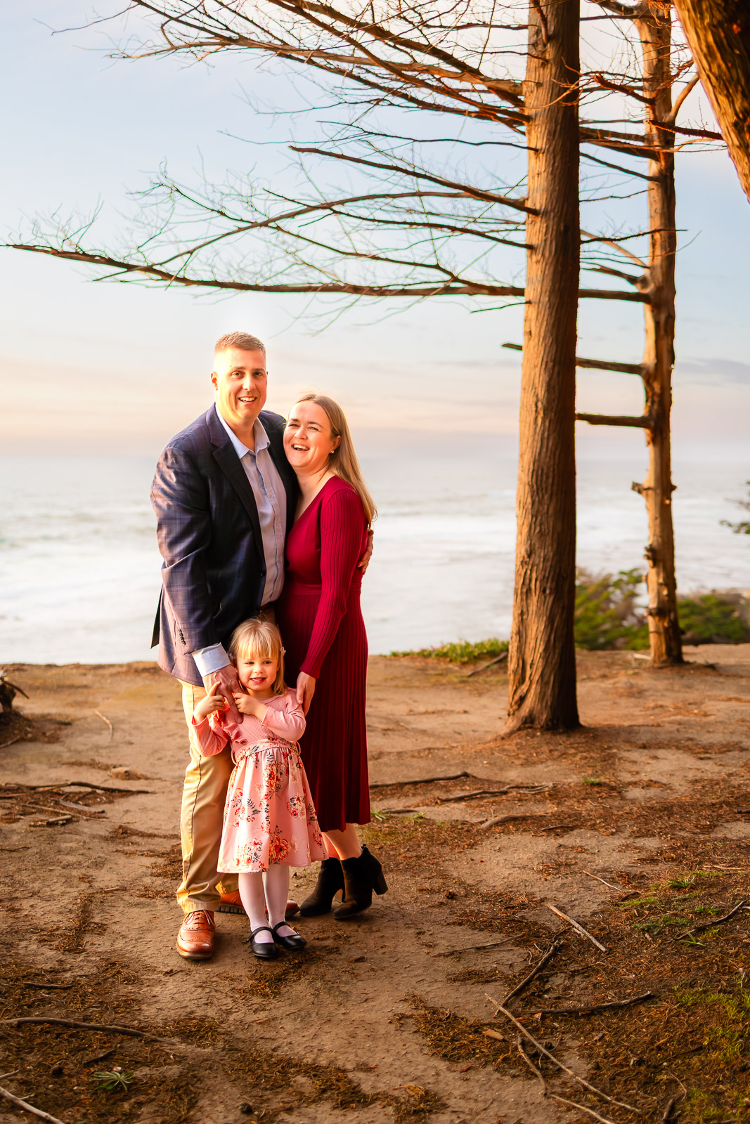 Family of three smiling during golden hour at a coastal cliffside with tall trees and ocean views in the San Francisco Peninsula.
