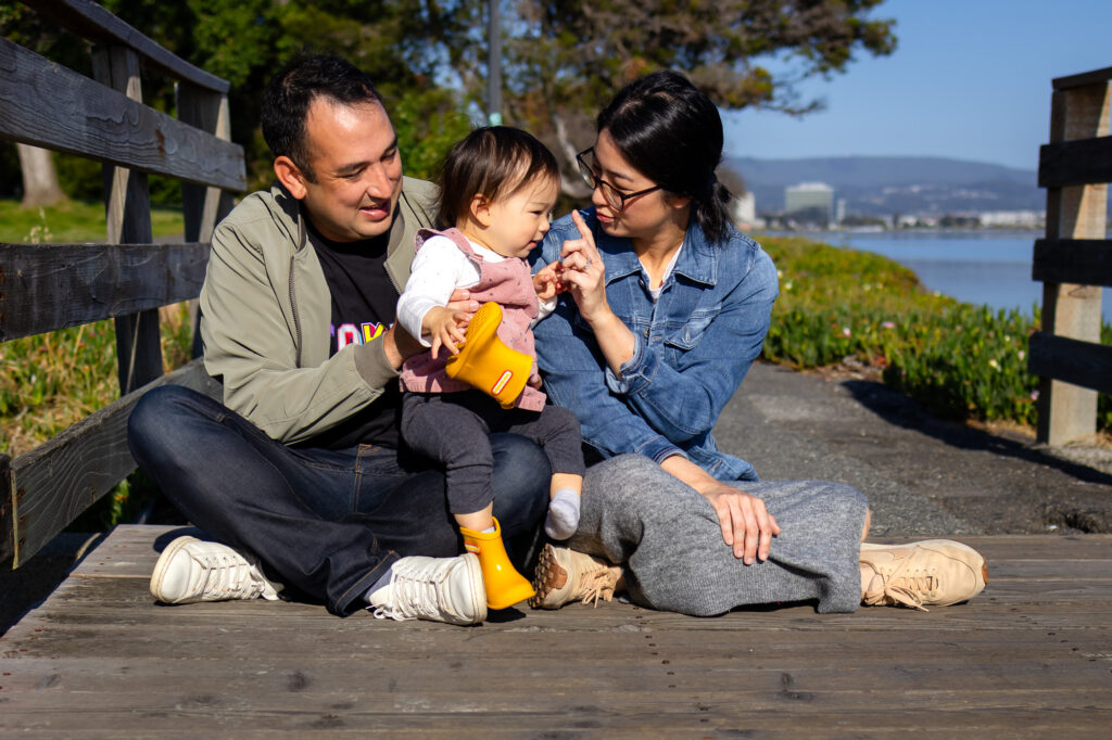 A mother and father sit close together on a Bay Trail wooden bridge while playing with their toddler during a relaxed outdoor Burlingame locations for family photos