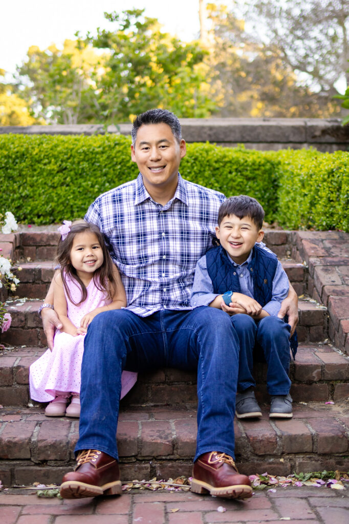 A father sits on brick steps with his two children, smiling and laughing together during a calm family portrait at one of the San Francisco Peninsula family photo locations in Burlingame
