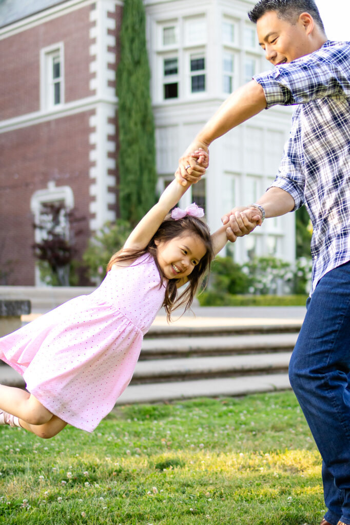 A father spins his young daughter by her hands as she laughs mid air during a playful family session at Burlingame family photo session locations.