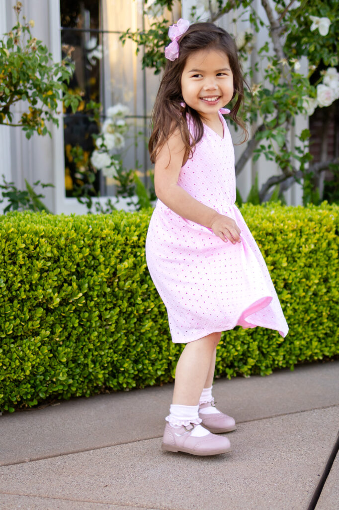 A young girl twirls and smiles while walking along a garden path surrounded by greenery and soft natural light.