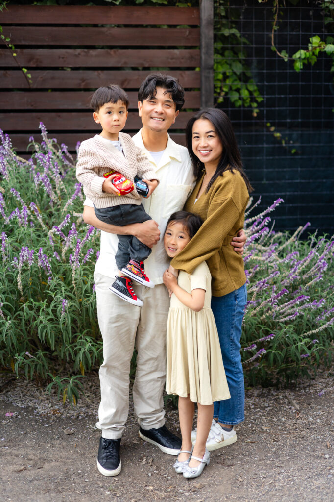 A family of four stands together among blooming flowers, holding one another closely during a relaxed outdoor session at photographer recommended family photo locations in Burlingame.