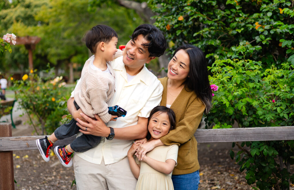 A family of four stands close together near a wooden fence, smiling and laughing as parents hold their young son while their daughter hugs in during a relaxed outdoor session.