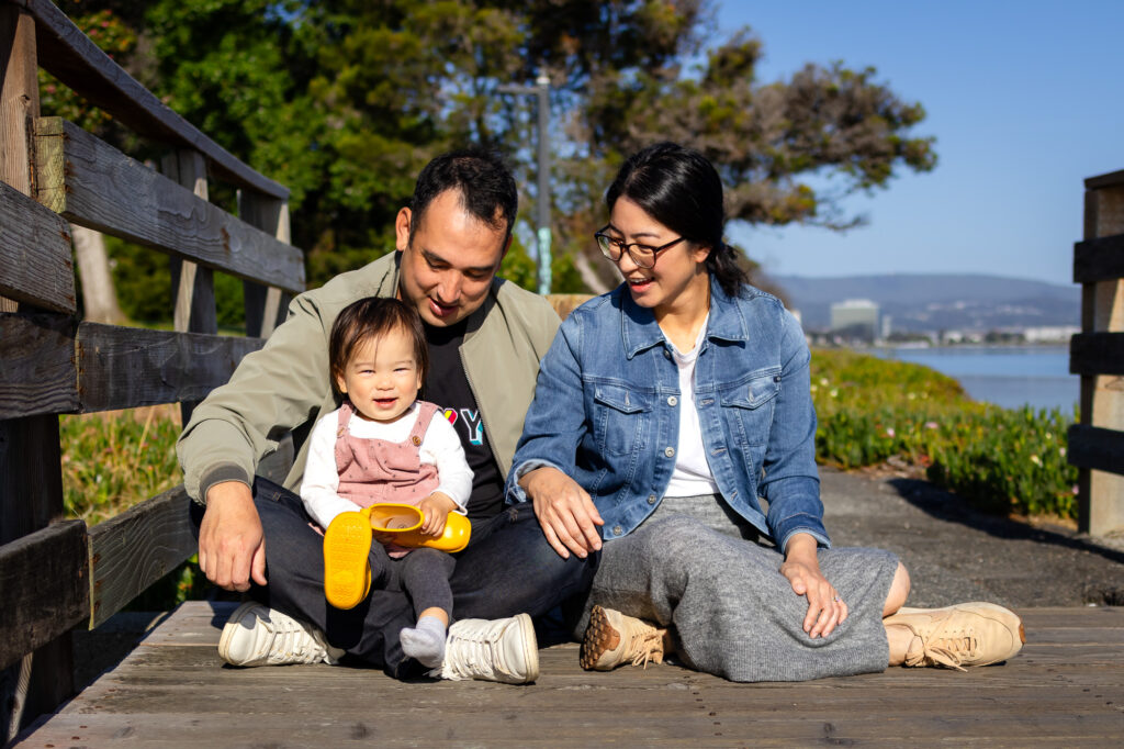 A toddler smiles while sitting between their parents on a wooden bridge, holding a yellow boot as the family at one of the best places for family photos in Burlingame