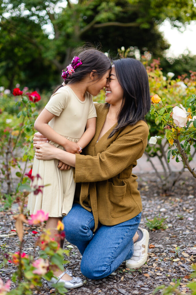 A mother and daughter lean in forehead to forehead, smiling and laughing together among blooming roses at one of the Burlingame Family Photo Locations.