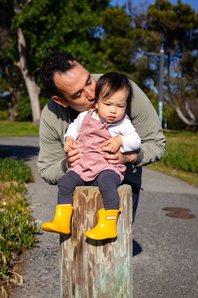 A father gently kisses his toddler’s cheek while the child sits on a wooden post along a quiet park path near the water on Bay Trail Burlingame