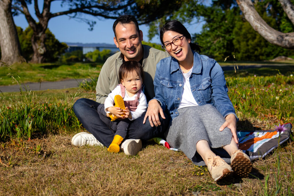 A family sits together on a picnic blanket in the grass with their toddler between them, smiling and enjoying the sunshine during a relaxed session at a Burlingame family photography locations