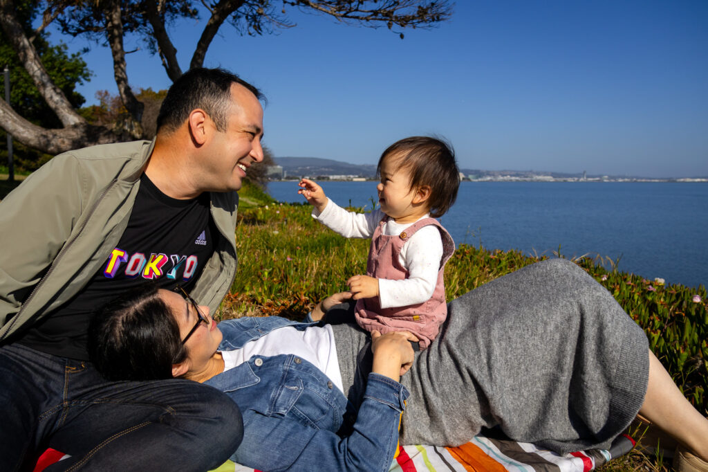 A toddler reaches toward their father while sitting on their mother’s lap on a picnic blanket near the water during a calm family photo session in Burlingame.