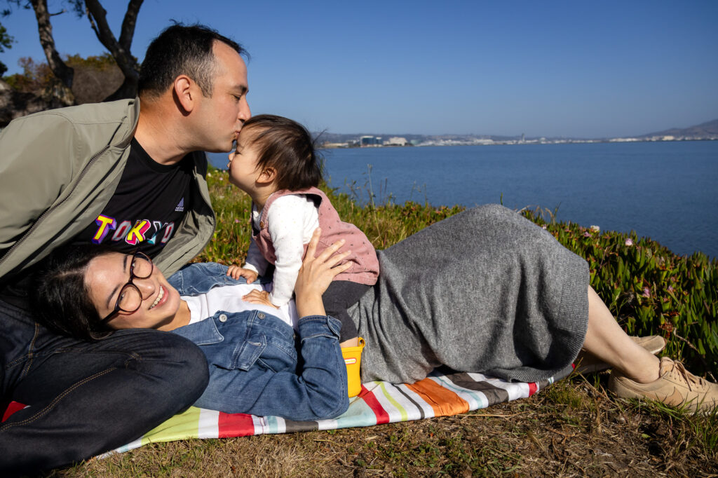 A father leans in to kiss his toddler while the child balances on their mother’s lap during a peaceful outdoor family portrait locations around Burlingame