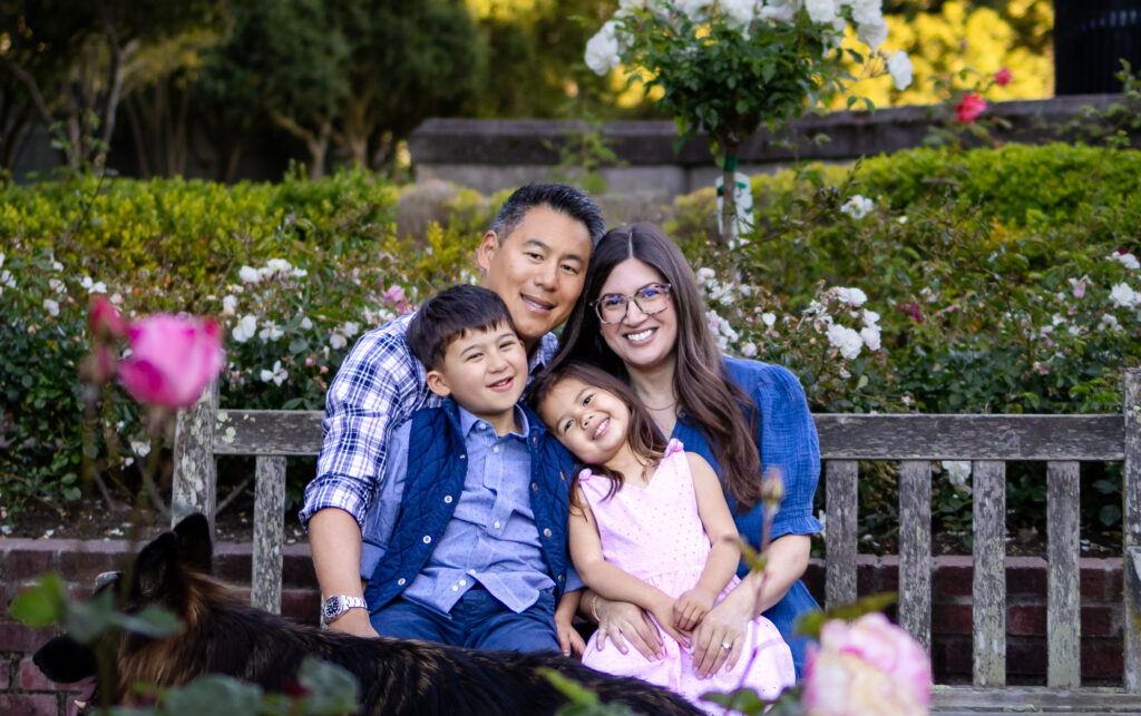 A family of four sits close together on a garden bench surrounded by blooming roses during an outdoor relaxed family photo settings in Burlingame