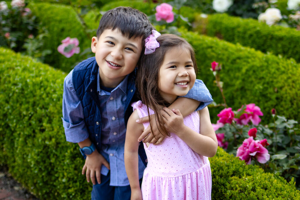 Two young siblings smile and hug while standing together in a garden filled with greenery and flowers during a family session in Burlingame.