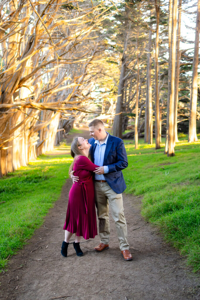 A couple stands close together on a dirt path beneath tall cypress trees, smiling at each other in warm golden light during their Half Moon Bay photography session.