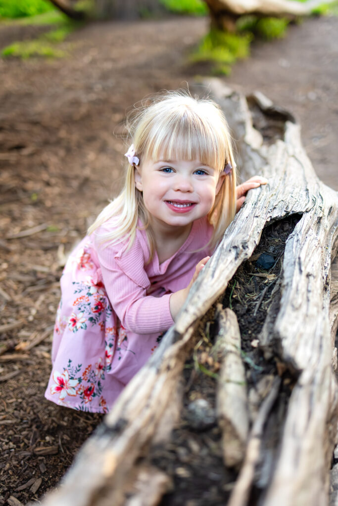 A young girl smiles at the camera while leaning on a weathered log, photographed by a family photographer.