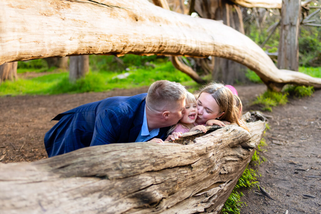 Both parents lean over a fallen tree to kiss their laughing daughter on each cheek, captured by a natural light photographer in Half Moon Bay.