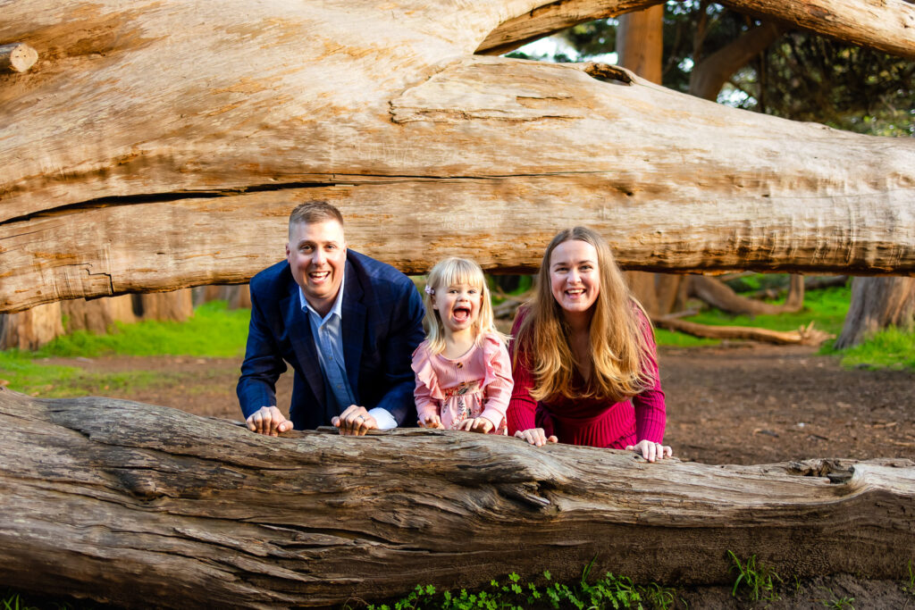 A family of three leans on a large fallen log, smiling and looking toward the camera during Half Moon Bay portrait session.