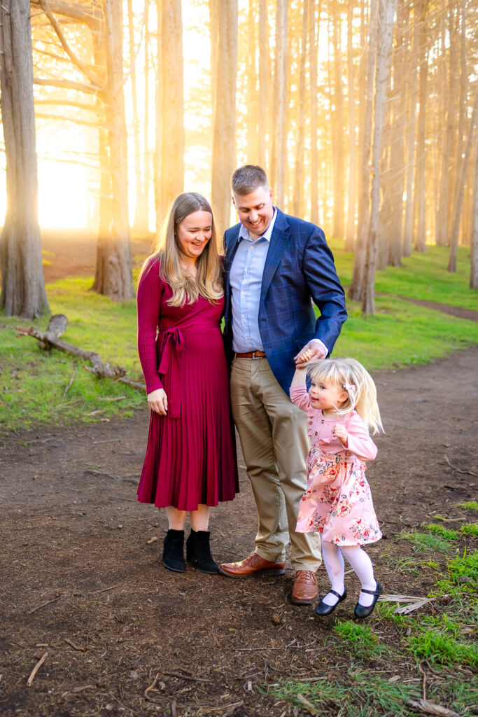 A father holds his daughter while the mother rests her hand on his chest as they stand among tall trees at sunset