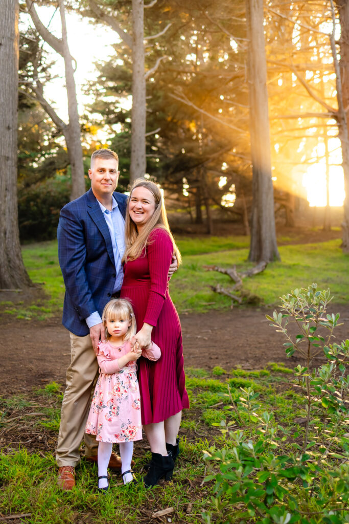 A mother and father stand close with their daughter between them, warm sunlight filtering through the trees during coastal family photography in Half Moon Bay.
