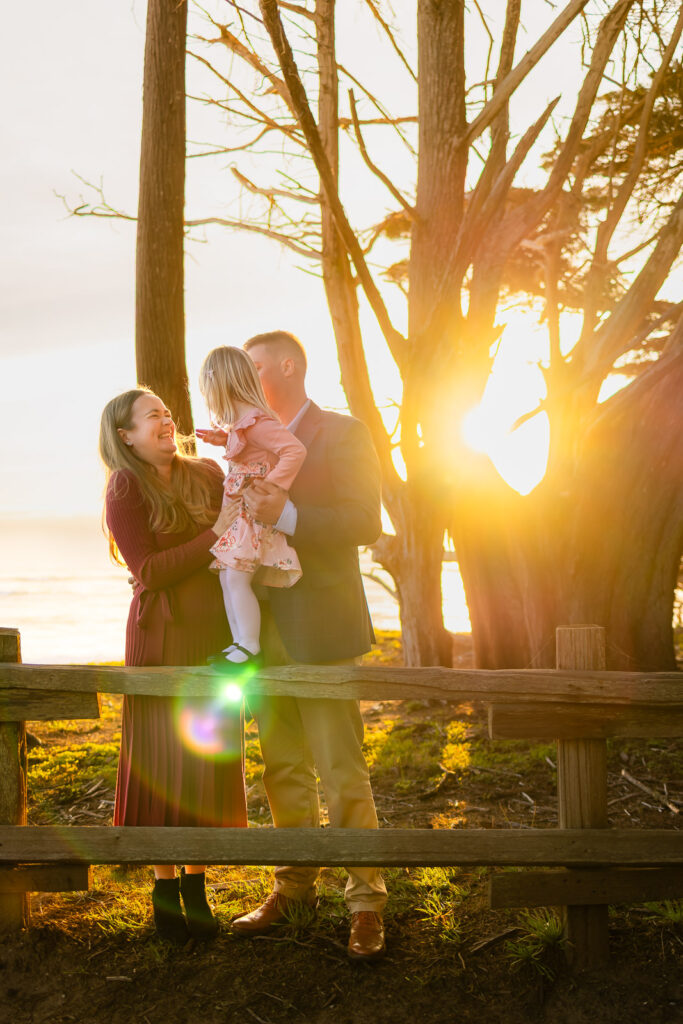 Parents lift their daughter while laughing near a wooden fence with the ocean glowing behind them, documented during beach sessions in Half Moon Bay.