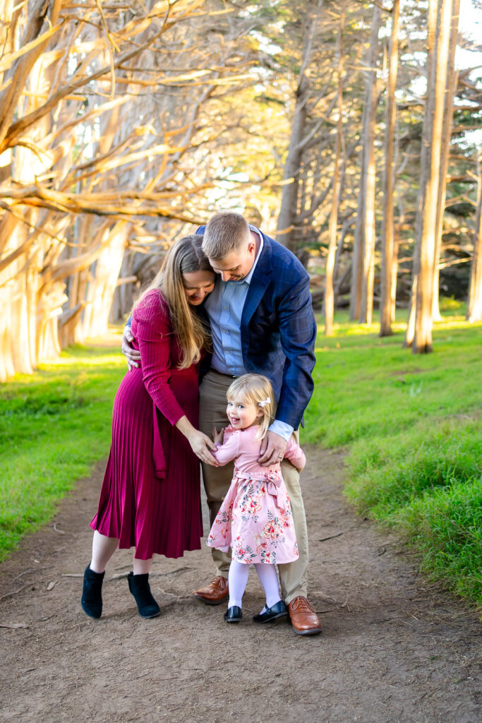 Parents lean down and hold their toddler daughter’s hands as she smiles on a wooded trail, captured by a photographer