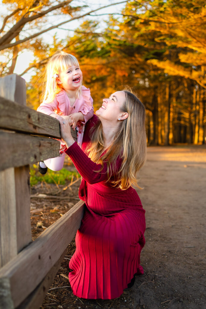 A mother kneels beside a wooden fence, lifting her laughing daughter as warm evening light filters through the trees, documented by a natural light photographer in Half Moon Bay.