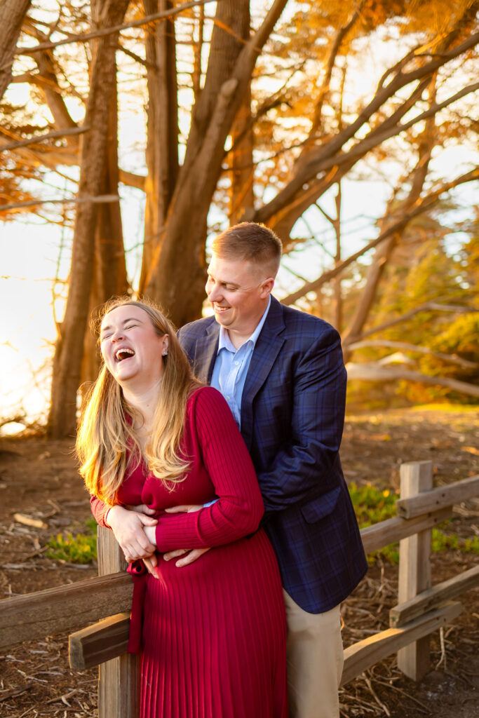 A husband embraces his wife from behind as she laughs with her head tilted back in the warm sunset glow, photographed by a Coastside photographer.