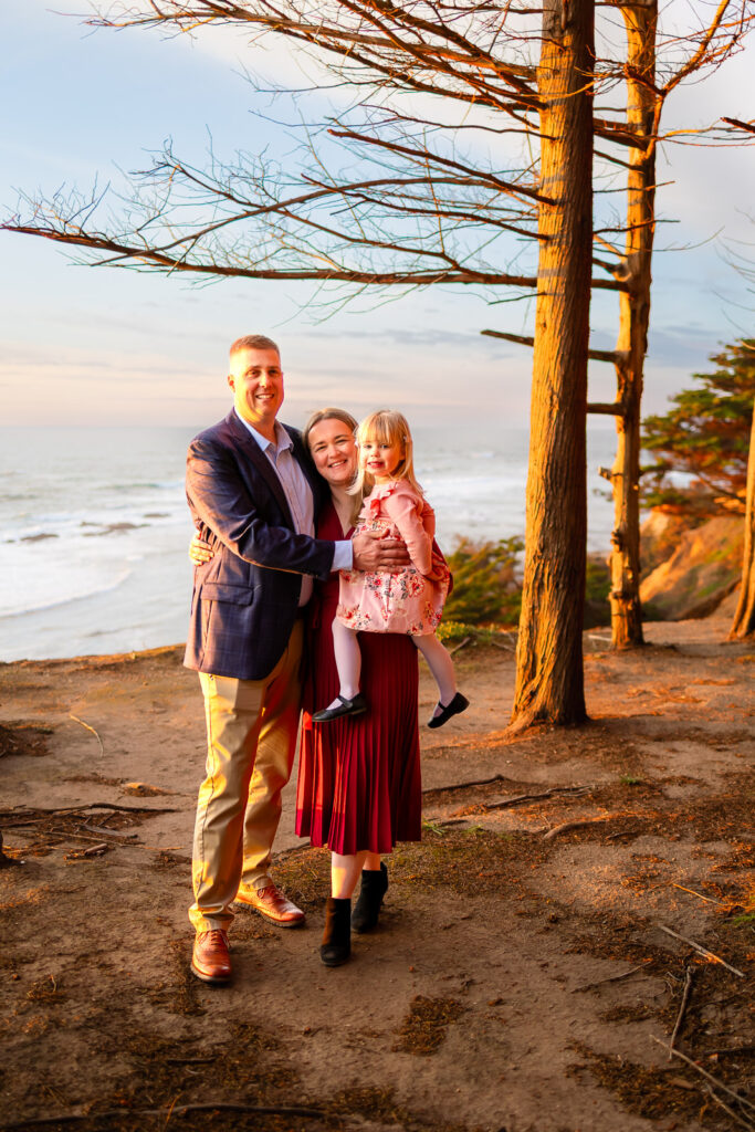 A family of three stands on a coastal overlook with the ocean behind them at sunset, photographed by a Half Moon Bay Photographer.