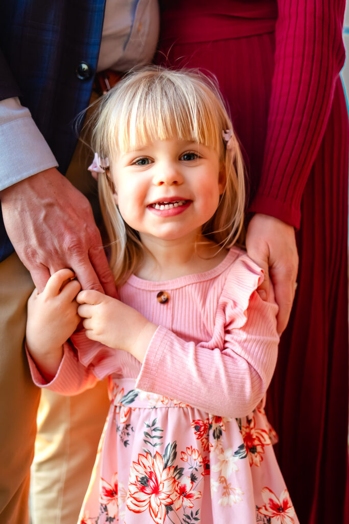 A close up portrait of a smiling toddler standing between her parents and holding their hands, captured by a Bay Area photographer serving Half Moon Bay.