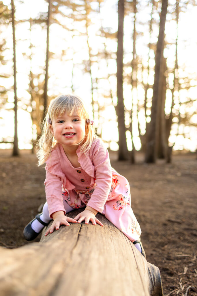 A young girl sits on a fallen log in the forest, smiling toward the camera during Half Moon Bay children photography at sunset.