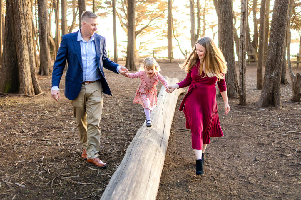 Parents hold their daughter’s hands as she carefully walks along a fallen tree trunk in the woods, photographed by an outdoor family photographer in Half Moon Bay.