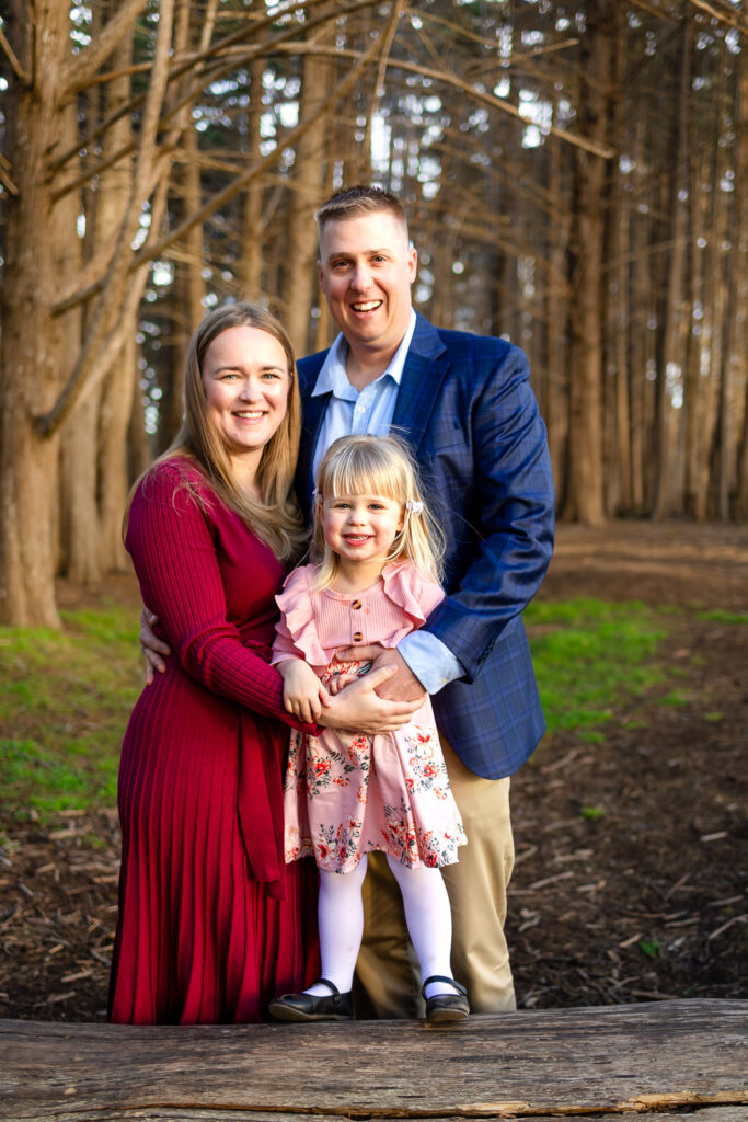 A mother and father stand close together holding their young daughter between them as they smile toward the camera in a cypress tree forest at golden hour, photographed by a Half Moon Bay Photographer.
