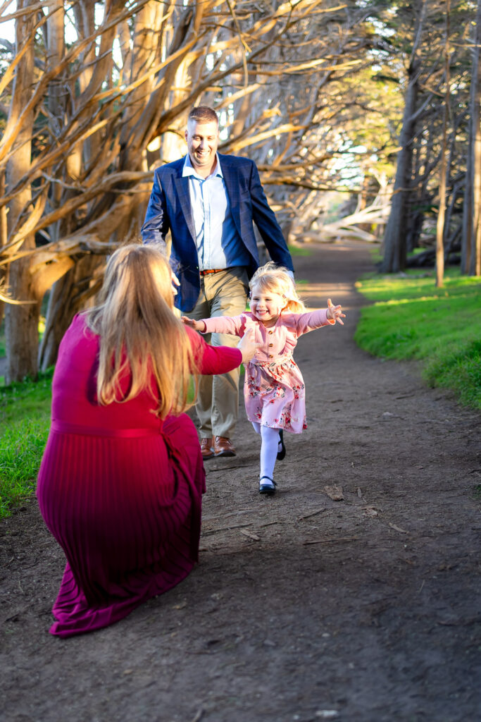 A little girl runs with open arms toward her kneeling mother on a wooded trail while her father walks behind them in warm evening light, captured by an outdoor family photographer in Half Moon Bay.