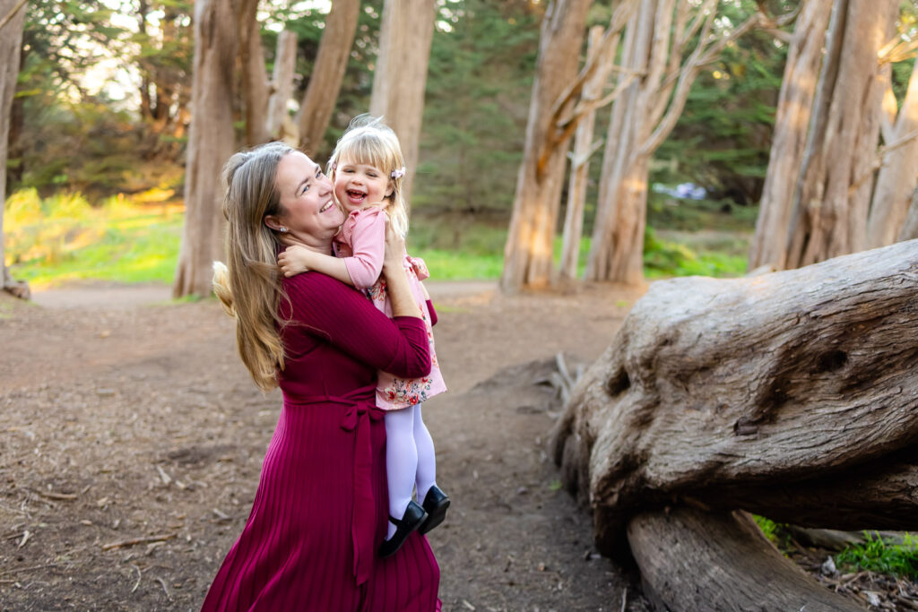 A mother holds her laughing toddler close near a large driftwood log, sharing a joyful hug during coastal sunset photography