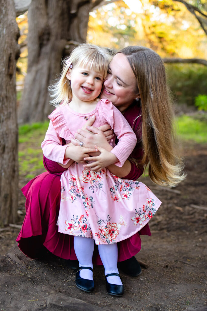 A mother kneels and kisses her daughter’s cheek as the little girl smiles toward the camera, documented by a Half Moon Bay maternity photographer.