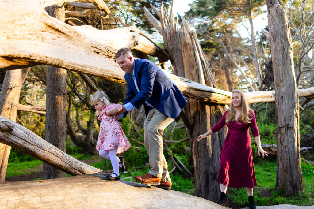 A father helps his toddler balance as she walks along a wide fallen tree while the mother follows behind, photographed by a Coastside photographer.