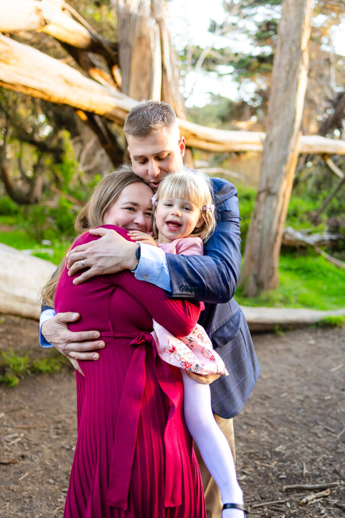 Parents embrace their daughter in a group hug as she beams with laughter, captured by a natural light photographer in Half Moon Bay.