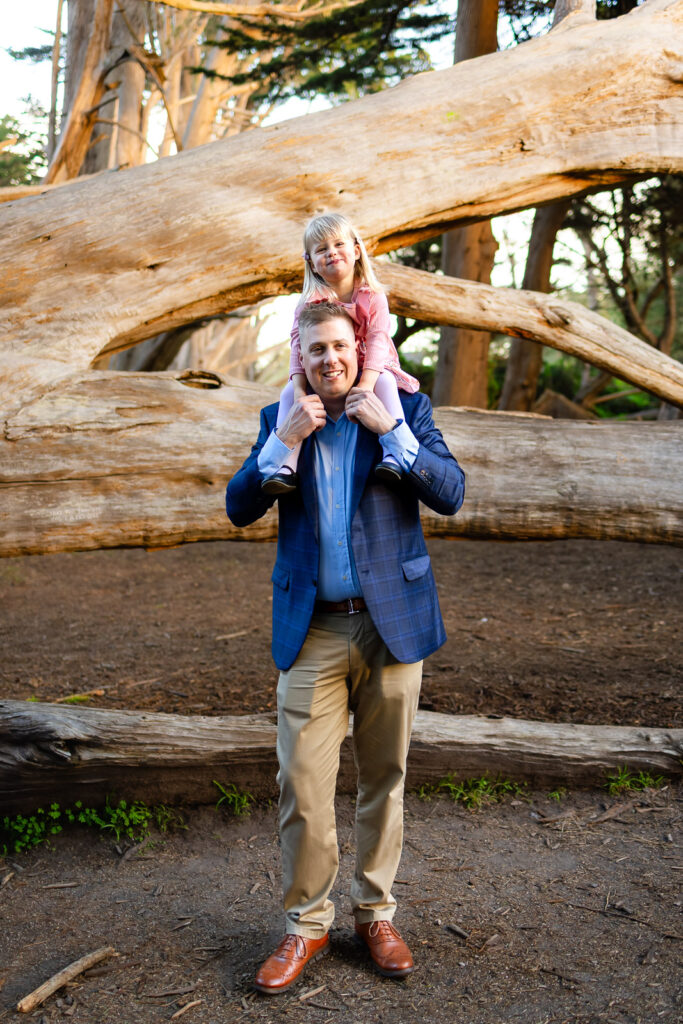 A father stands with his daughter sitting on his shoulders beneath layered driftwood logs, photographed by a Peninsula photographer for Half Moon Bay families.