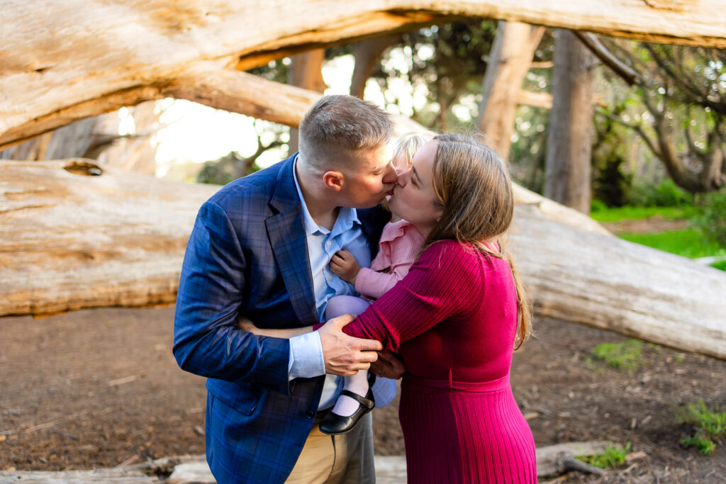 A family of three stands together in front of large fallen cypress logs, kissing during a session with a Half Moon Bay Photographer.