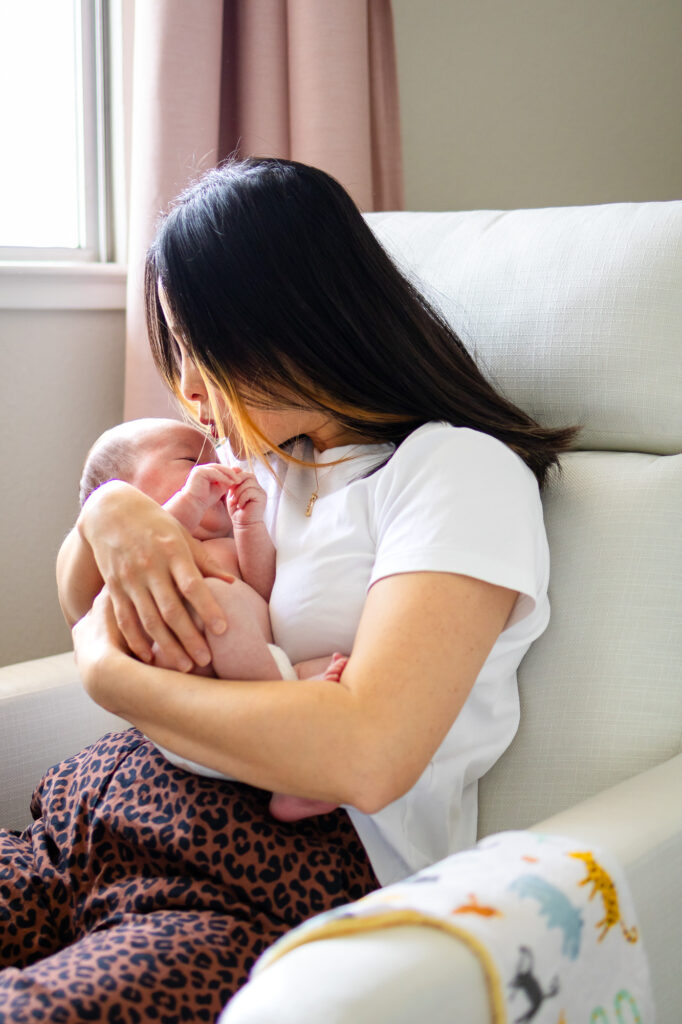 A mother sits in a light upholstered chair near a window, gently cradling her newborn against her chest during an intimate In-Home Newborn Photography for First-Time Parents session.