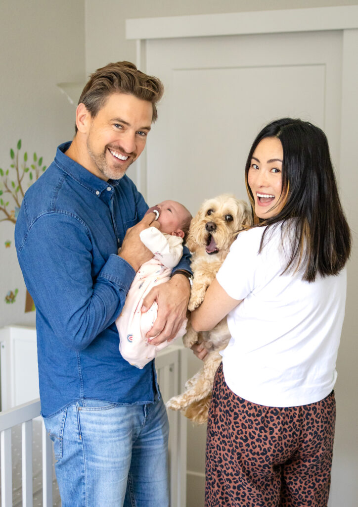 A father holds his newborn while standing beside the mother who cradles their small dog, both parents smiling toward the camera in a bright nursery with white walls.